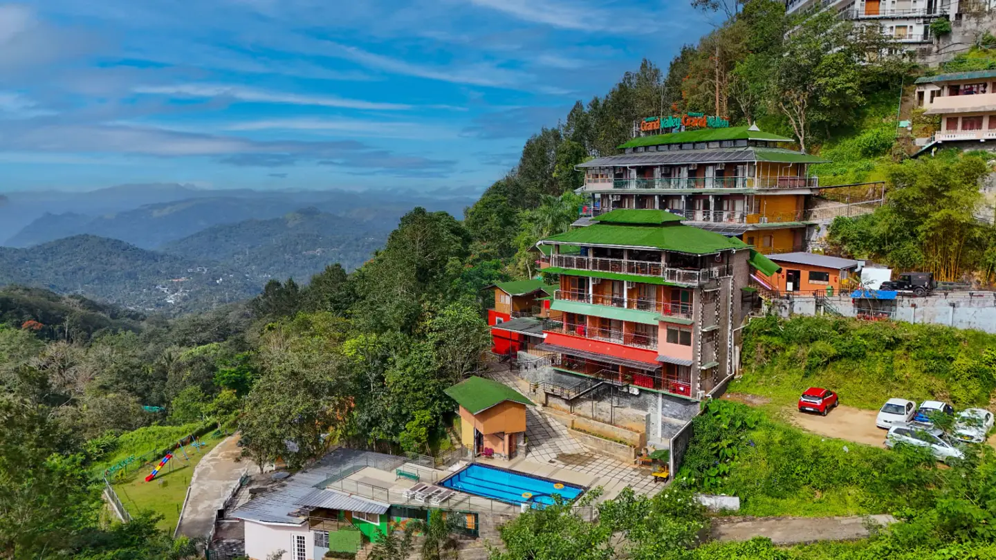 Aerial view of Grand Valley Resort's green-roofed blocks and pool set into the misty tea hillside at 2nd Mile, Munnar